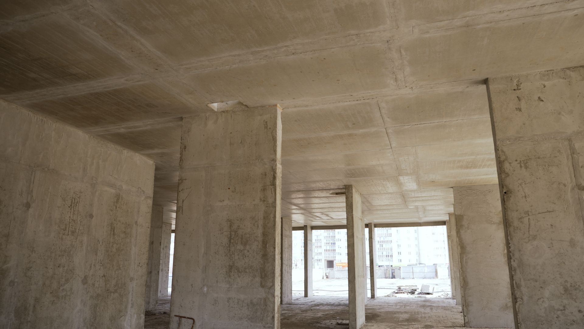 Intérieur en béton brut, vide, avec colonnes et grandes ouvertures vitrées.