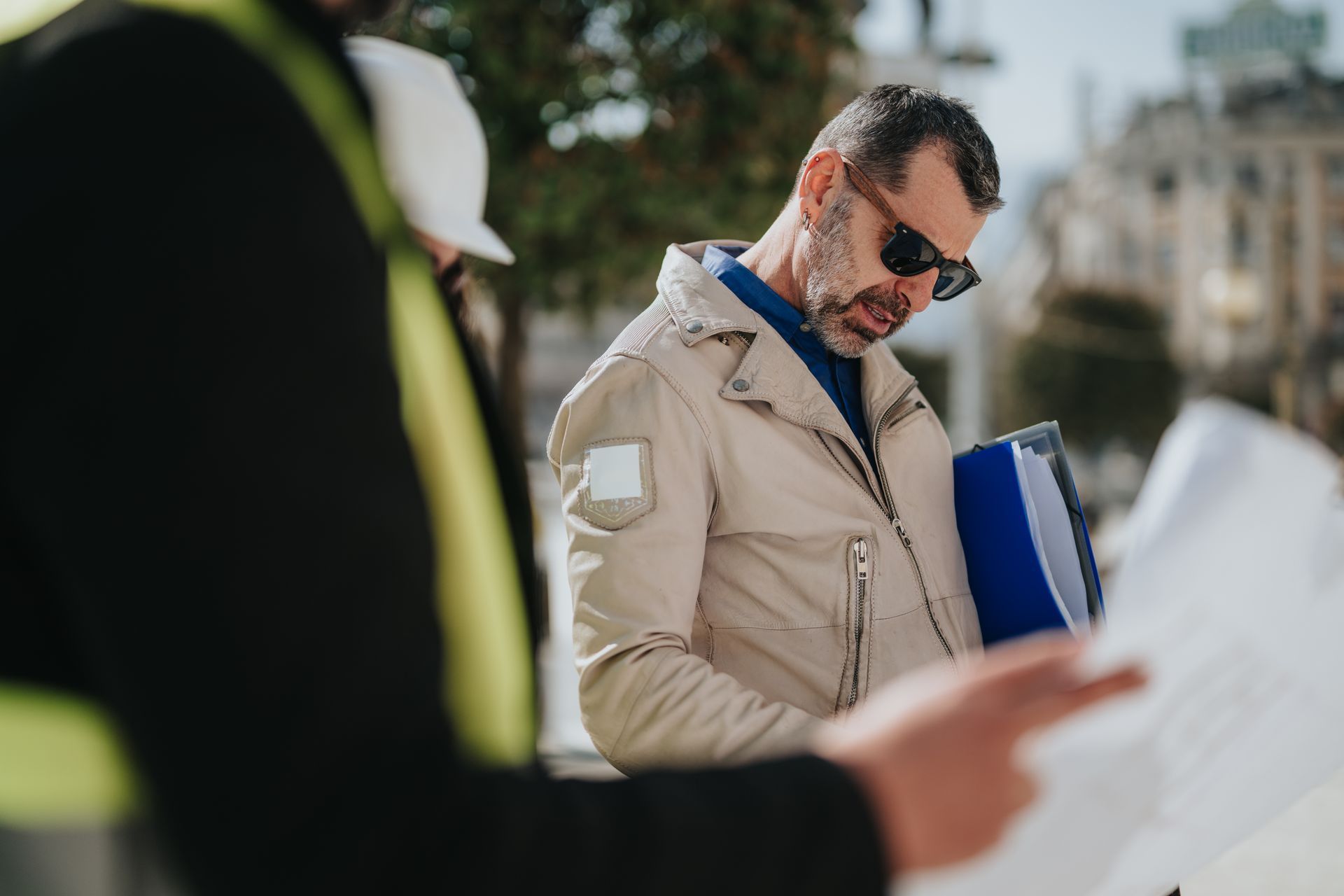 Un homme en manteau beige et lunettes de soleil, tenant un dossier bleu à l'extérieur, regarde des papiers.