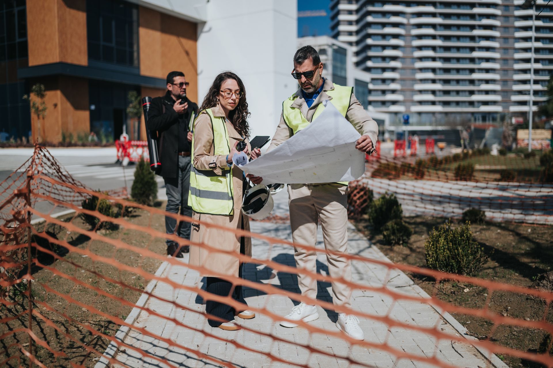 Des ouvriers examinent une carte sur un chantier de construction, derrière une clôture de sécurité orange.