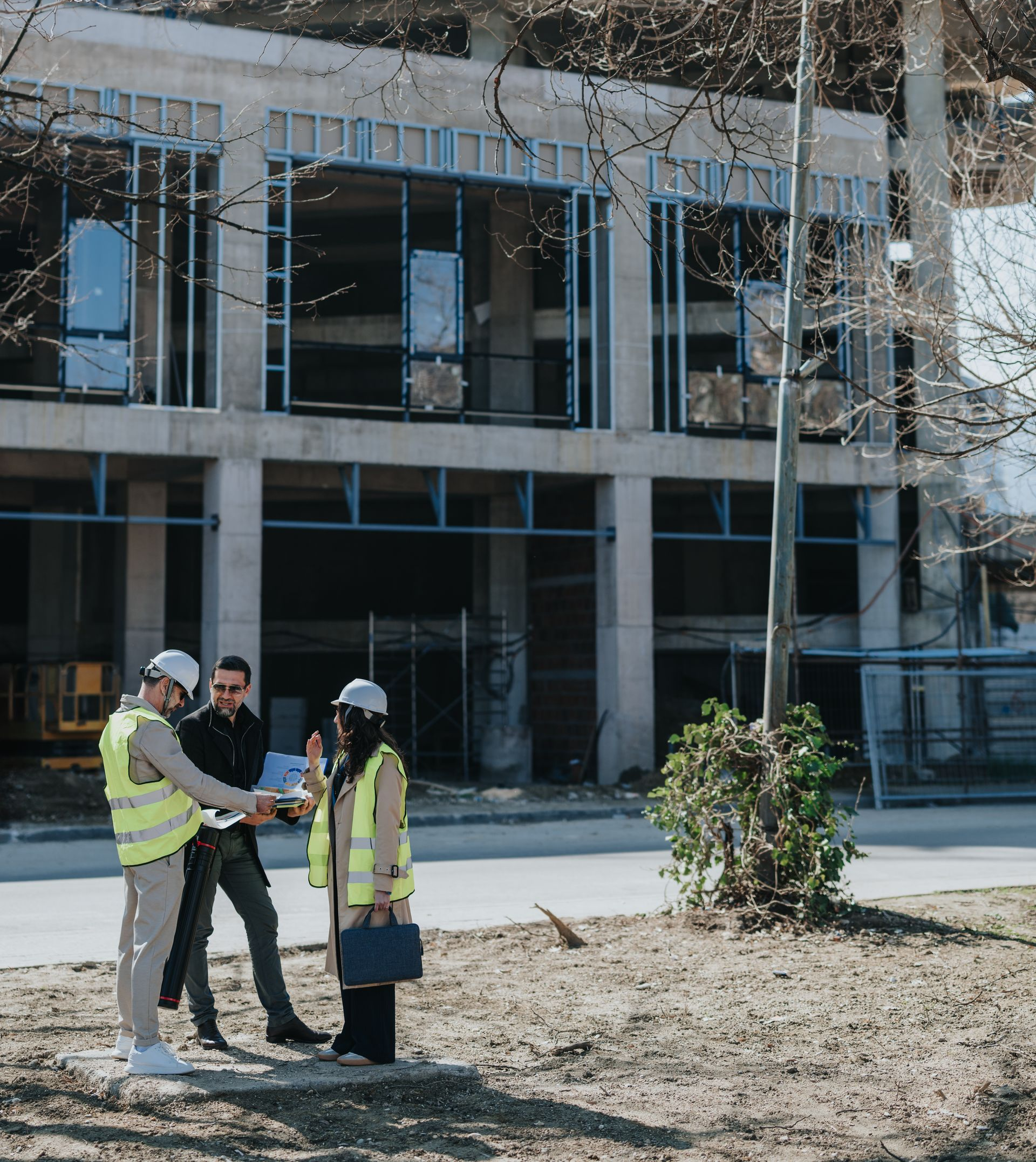 Trois ouvriers portant des gilets de sécurité et des casques de chantier discutent devant un bâtiment en construction.