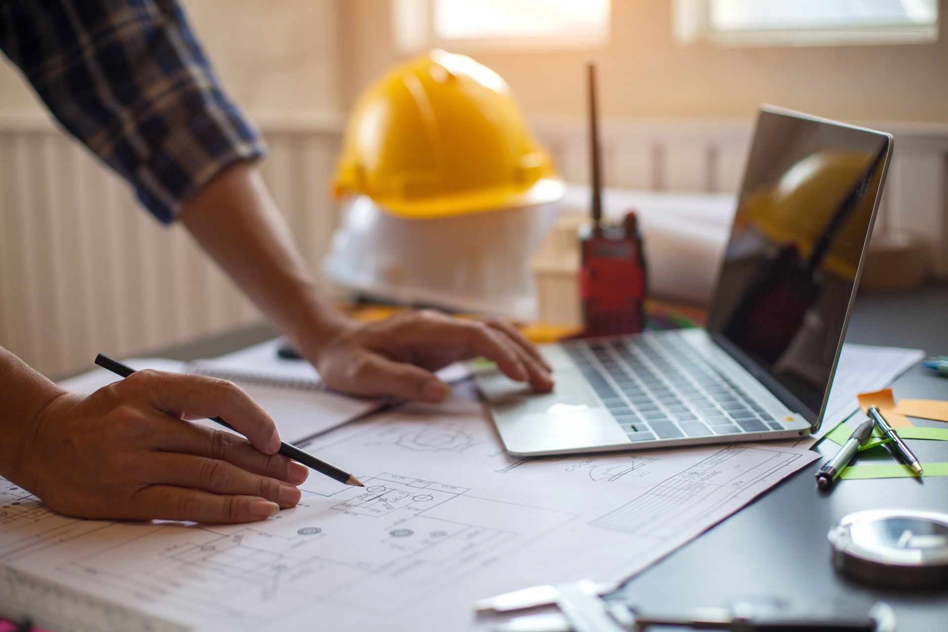 Des mains dessinent des plans à côté d'un ordinateur portable et d'un casque de chantier jaune sur une table de travail