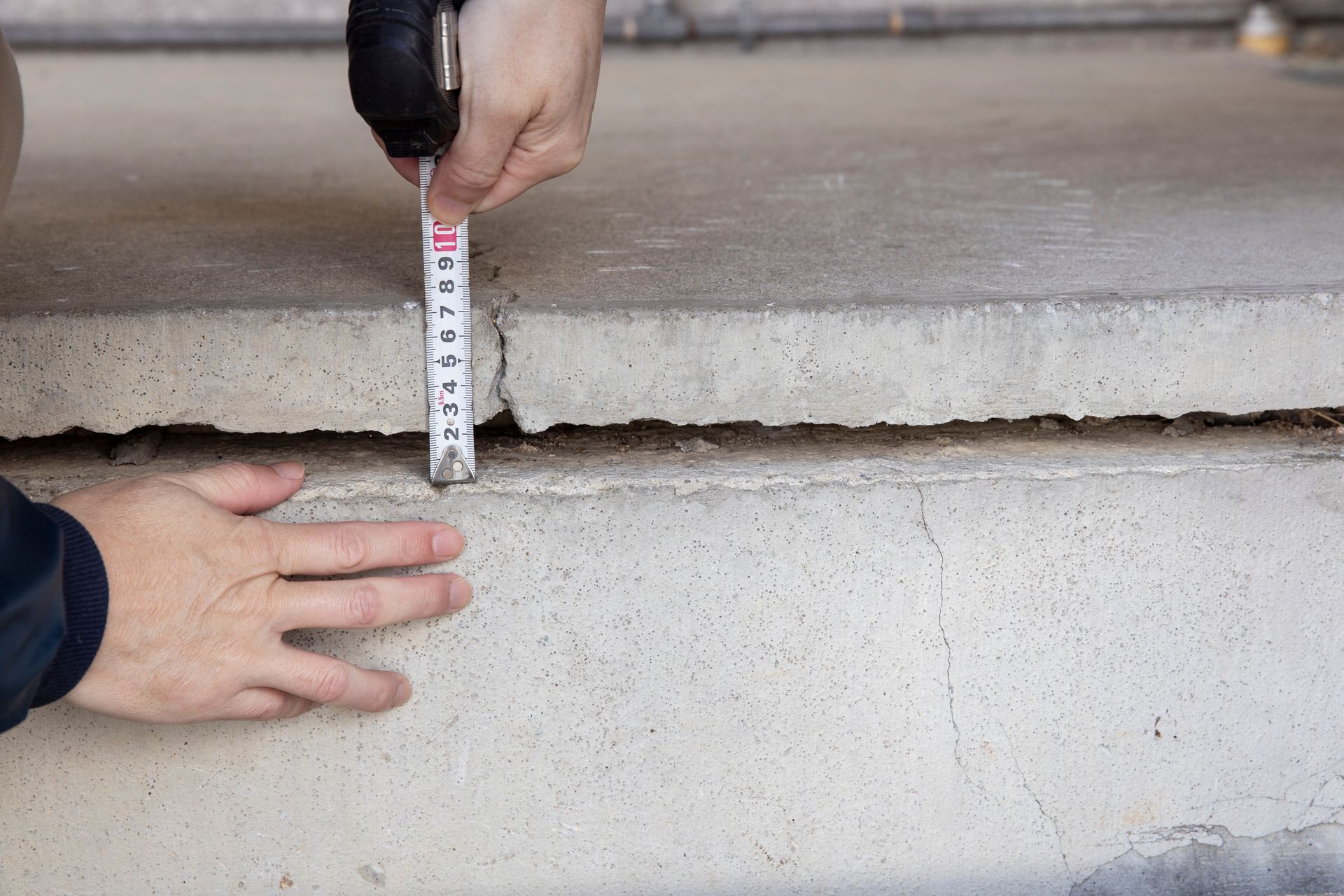 Des mains mesurent une fissure dans une dalle de béton à l'aide d'un mètre ruban, à l'extérieur.