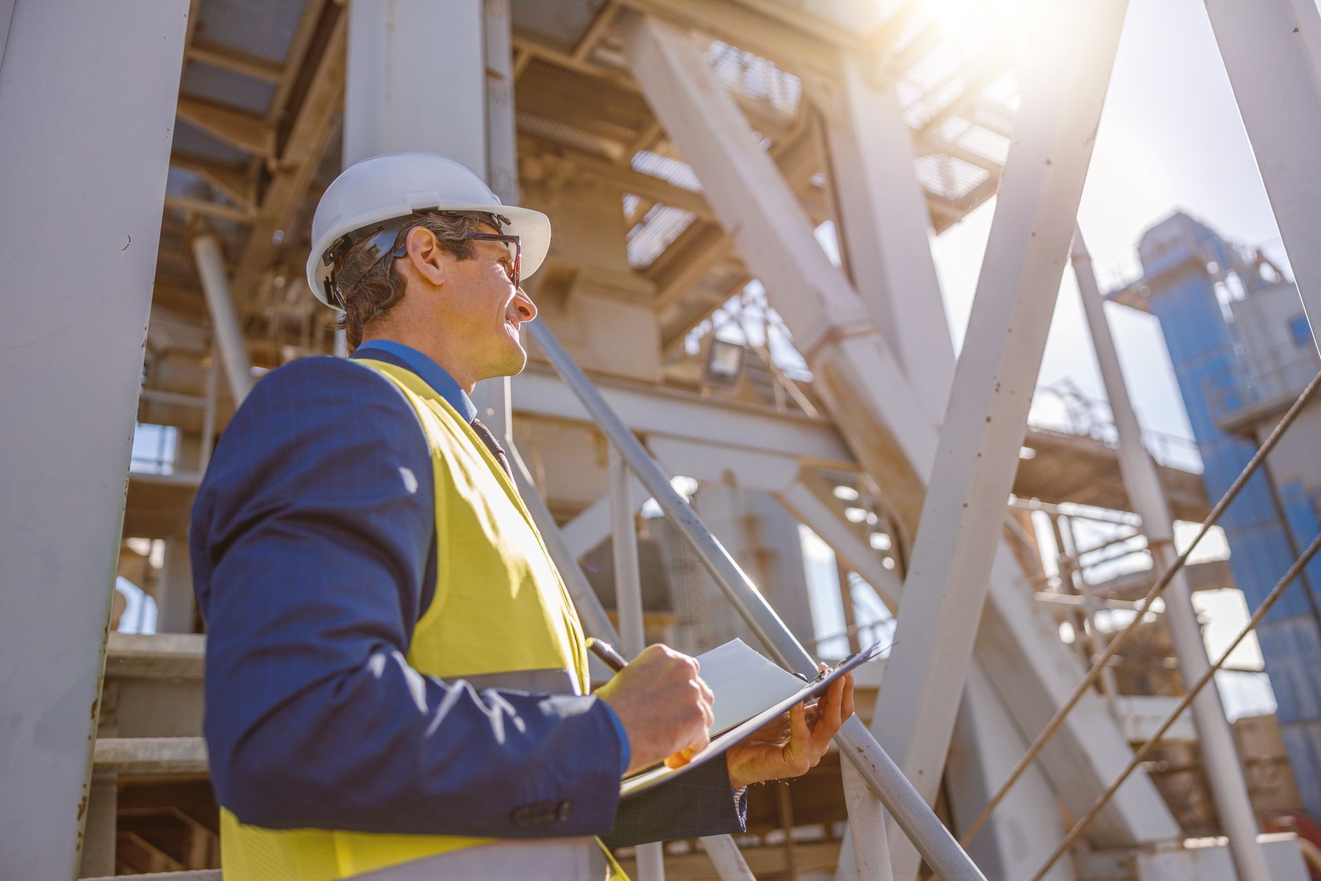 Un ingénieur portant un casque et un gilet jaune inspecte une structure métallique industrielle en plein soleil.