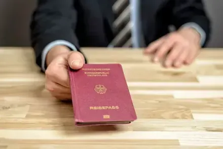 Persona con traje extendiendo un pasaporte color burdeos sobre una mesa de madera.