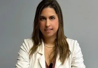 Mujer con cabello largo y castaño, vestida con un blazer blanco, sonriendo a la cámara sobre un fondo gris.