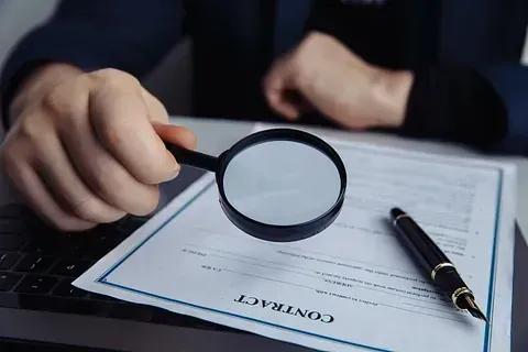 Una persona examina un contrato con una lupa; junto a ella hay un bolígrafo.