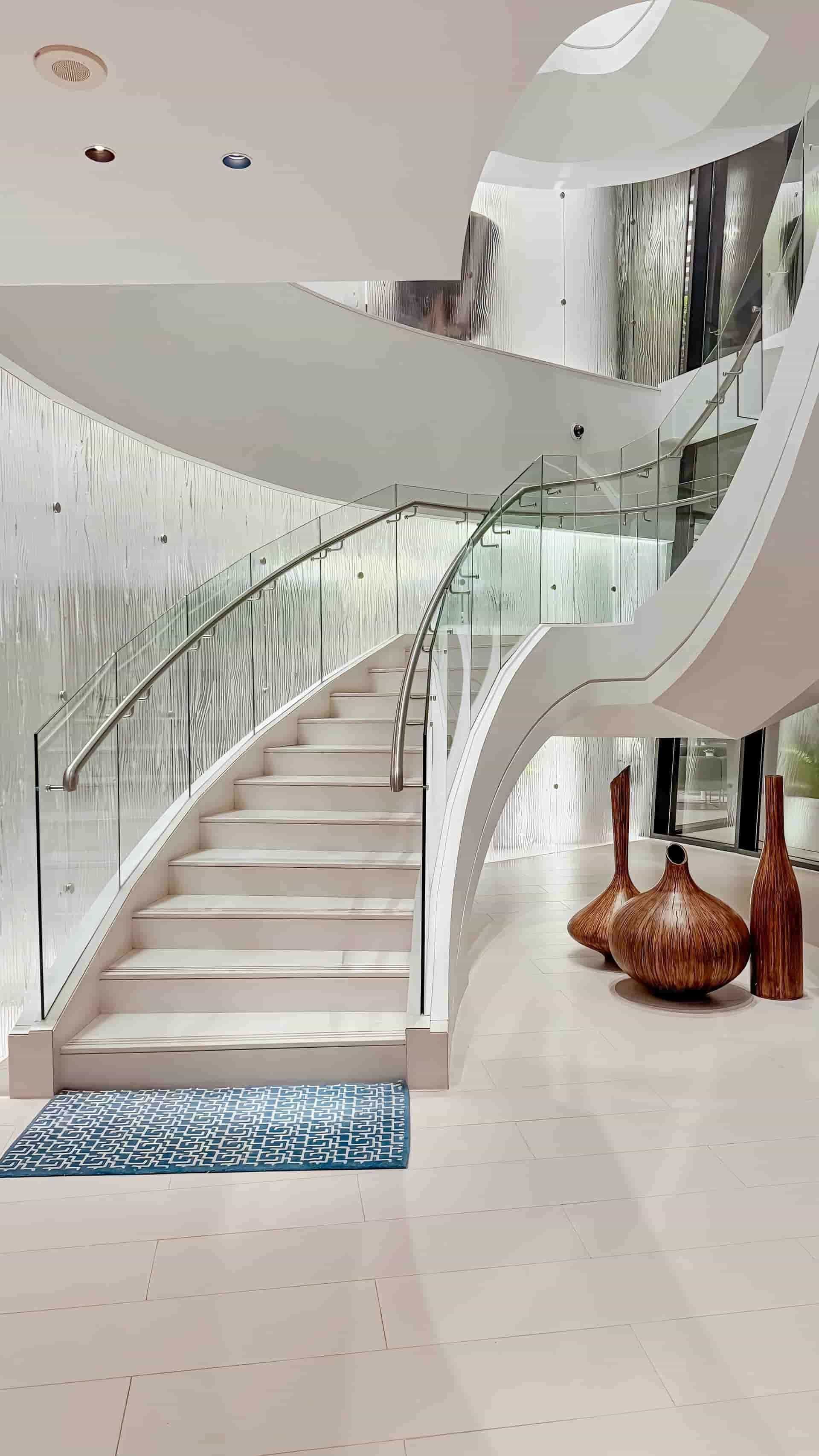 Escalera moderna blanca con barandillas de cristal. Jarrones decorativos se encuentran cerca, sobre el suelo de color claro.