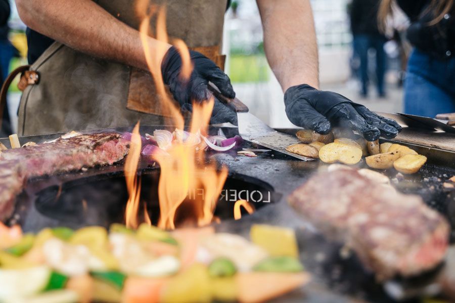 Koch grillt Fleisch und Gemüse auf einem Flachgrill; Flammen sichtbar, trägt Handschuhe.