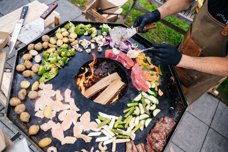 Person grillt Gemüse und Fleisch auf einem großen Außengrill mit einer zentralen Feuerstelle.