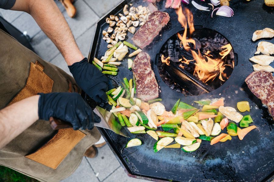 Person grillt Gemüse und Steak auf einem runden Grill mit offener Flamme.