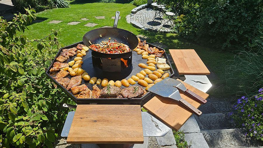 Outdoor-Kochsetup mit Fleisch, Gemüse und Kartoffeln auf einem großen, achteckigen Grill mit Holzschneidebrettern.
