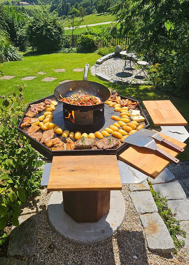 Kochgelegenheit im Freien mit Essen auf einem großen Grill, umgeben von Kartoffeln und Fleisch.