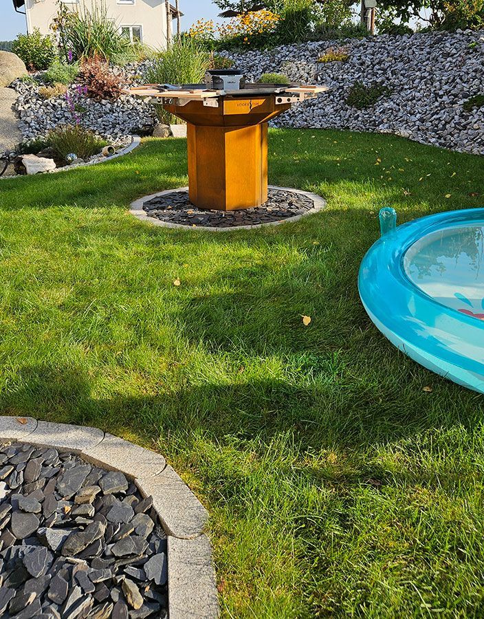 Ein Hinterhof mit einem Grilltisch auf einer Rasenfläche, einem kleinen Pool und einem Steingarten im Hintergrund.