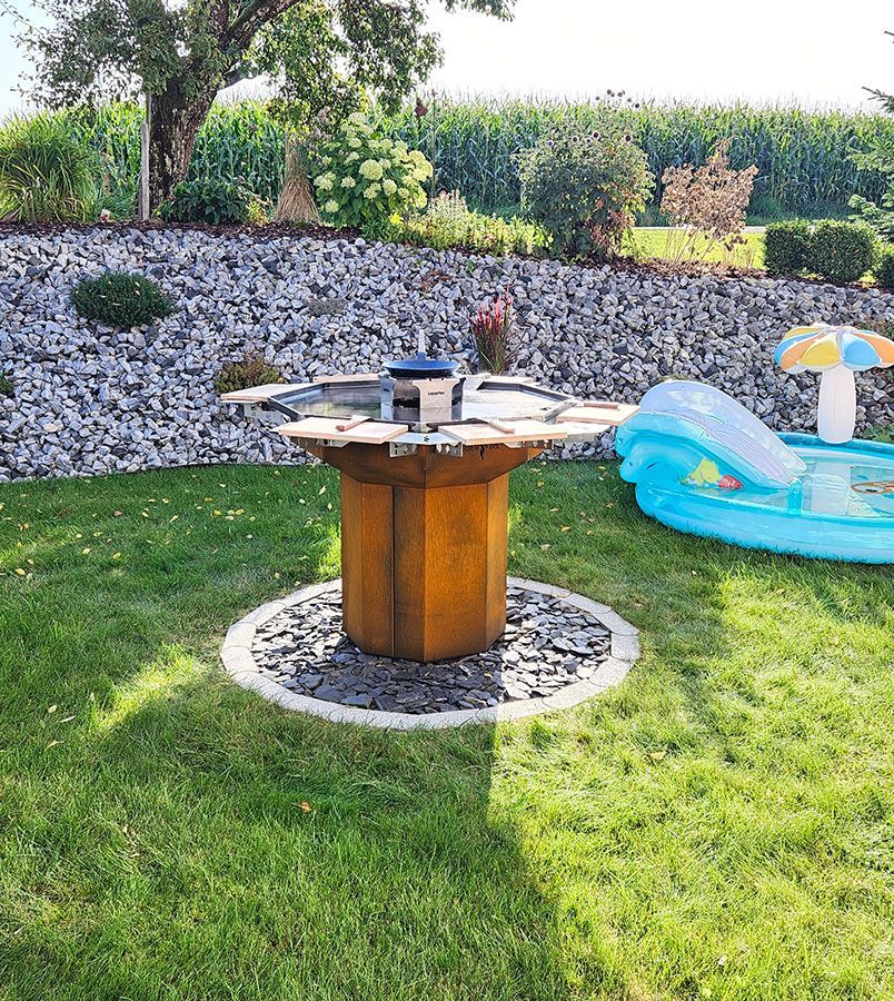 Rostiger Grilltisch aus Metall im Freien in einem grasbewachsenen Garten mit Steinhintergrund, kleiner Pool.