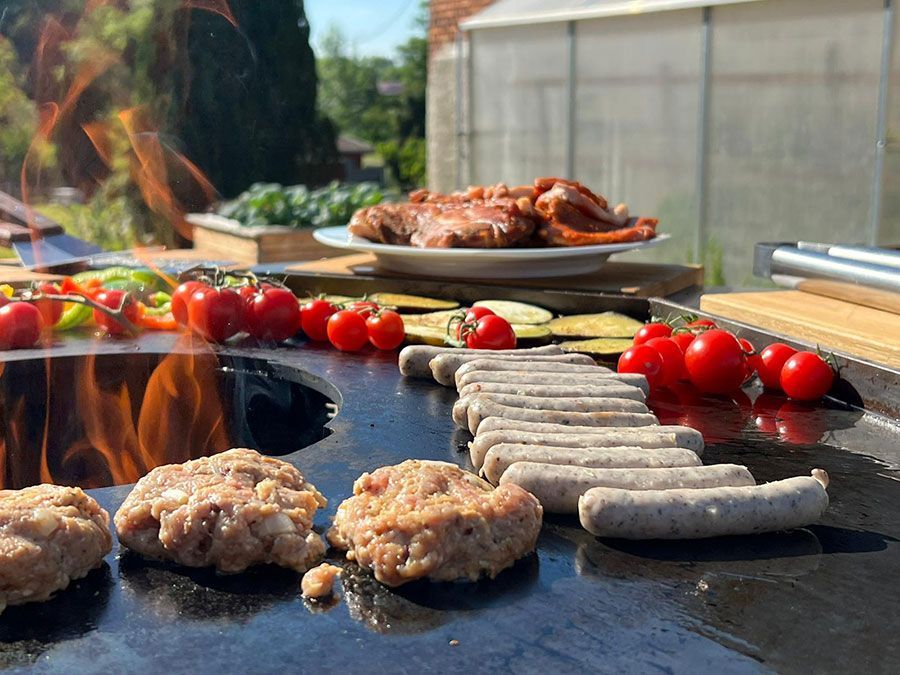 Grillen im Freien: Burger, Würstchen, Tomaten, Paprika, Zucchini und ein Teller mit gegartem Fleisch liegen auf dem heißen Grill.