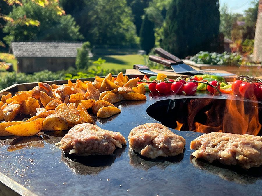 Außengrill mit Kartoffeln, Burgern und Gemüse, die über Flammen gegart werden.