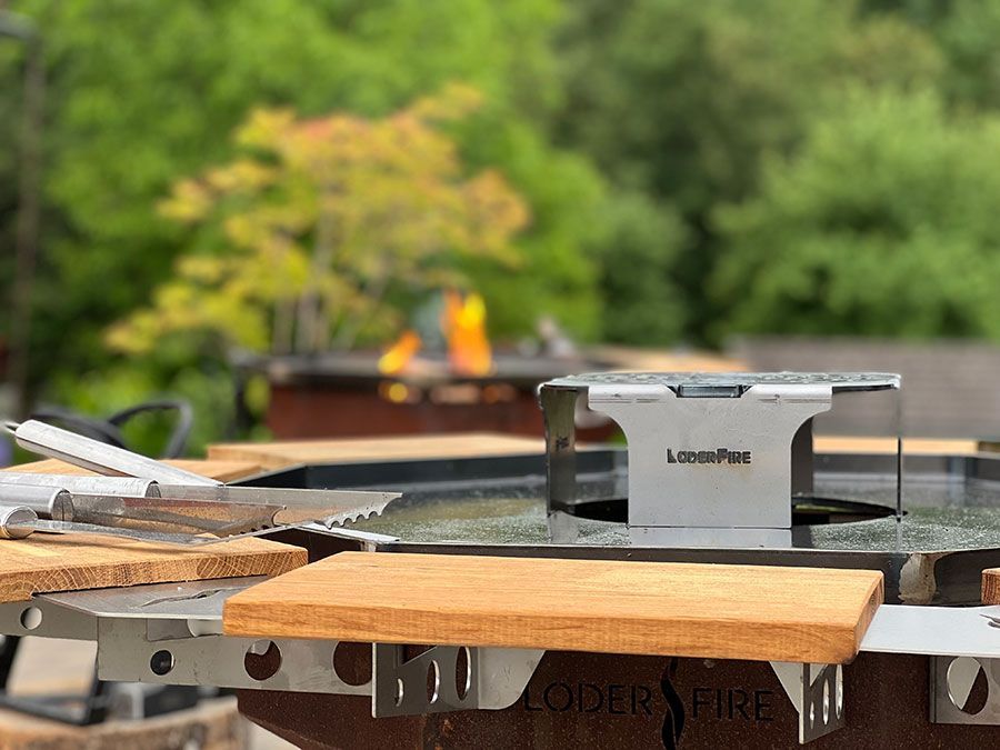 Nahaufnahme einer Feuerstelle aus Edelstahl mit Holzakzenten, unscharfer Hintergrund eines Hinterhofs mit brennendem Feuer.