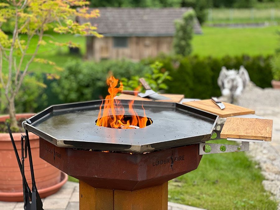 Eine Feuerstelle mit Flammen, umgeben von einer Kochfläche und hölzernen Vorbereitungsbrettern; im Freien.