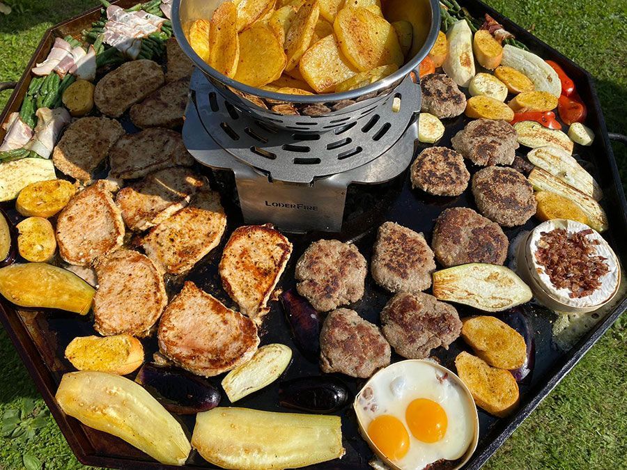 Eine Auswahl an Grillgerichten, angerichtet auf einer Grillplatte mit einem zentralen Frittierkorb; darunter Brot, Eier, Frikadellen und Gemüse.