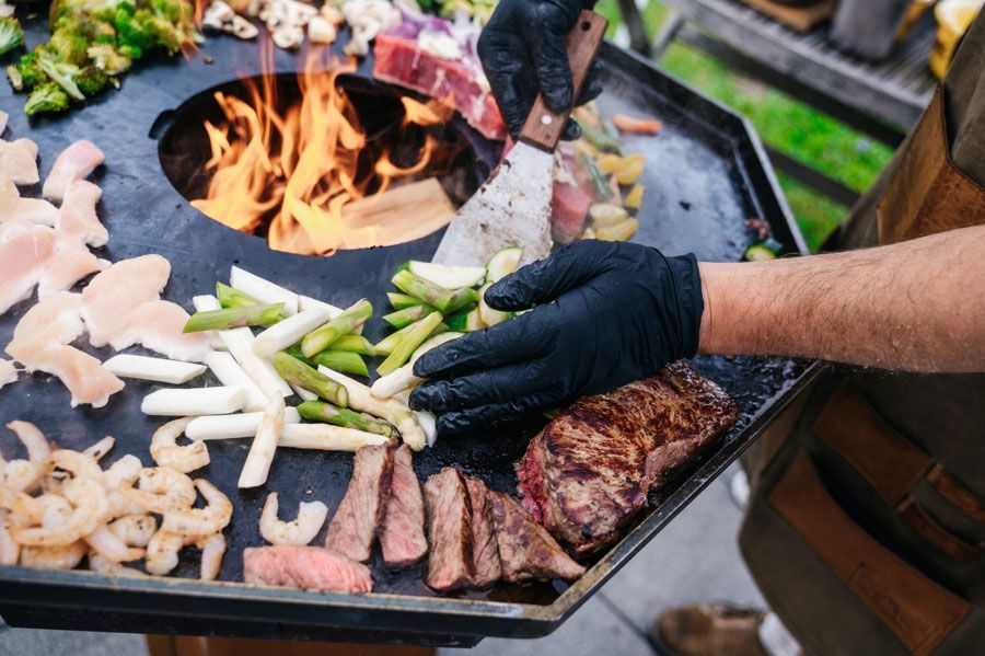 Der Koch grillt verschiedene Lebensmittel, darunter Fleisch und Gemüse, auf einem Flachgrill mit Flammen in der Mitte.