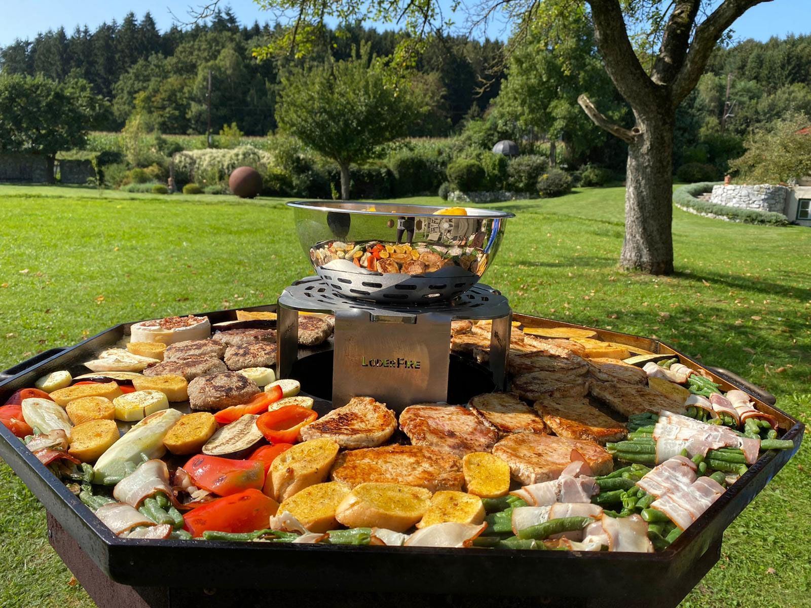 A grill loaded with various cooked food, on a green lawn with trees in the background.