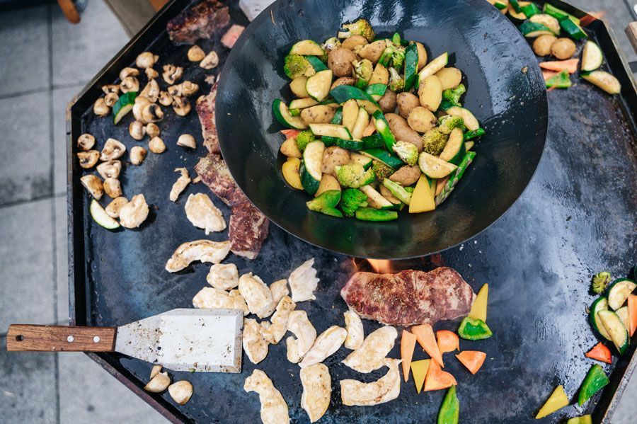 Außengrill mit Gemüse, Hühnchen und Steak auf einer großen, flachen Oberfläche; Wok in der Mitte.