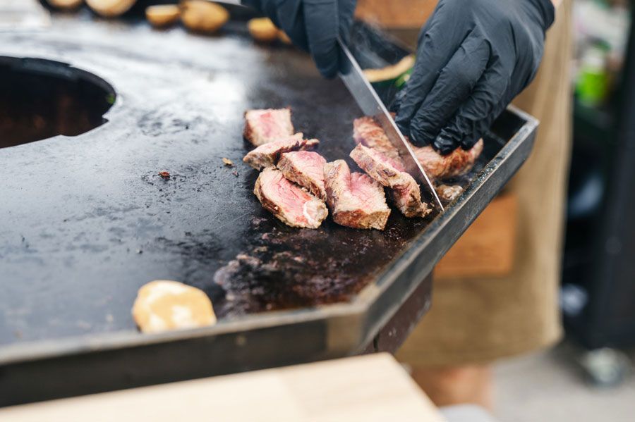 Koch mit schwarzen Handschuhen schneidet gekochtes Steak auf einer Grillplatte.