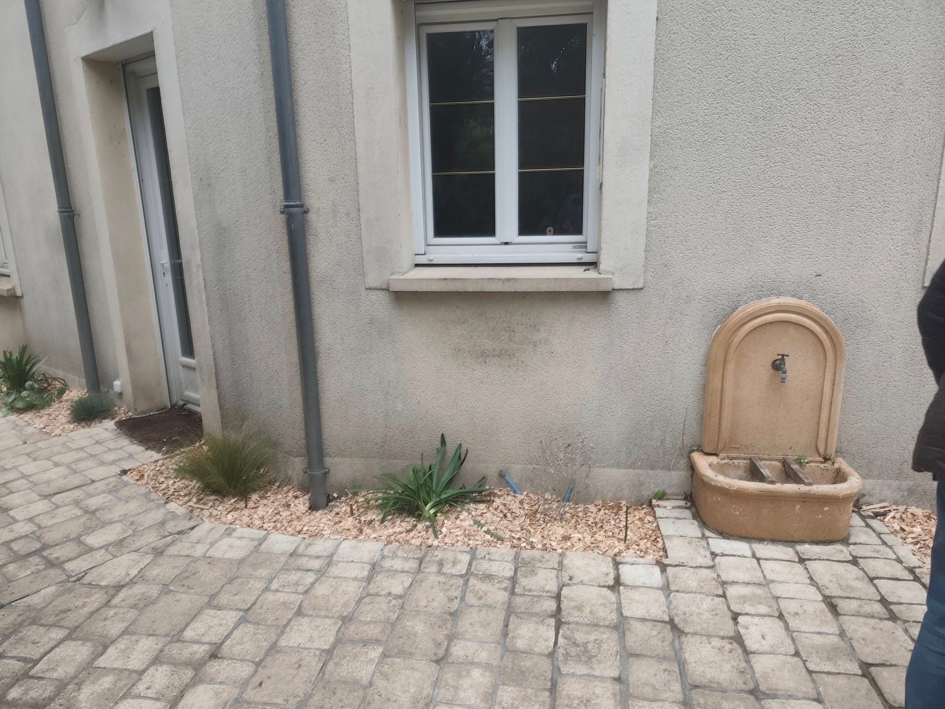 Une fontaine en pierre est adossée à un mur de maison en stuc, à côté d'une fenêtre, avec une terrasse pavée et de petites plantes.