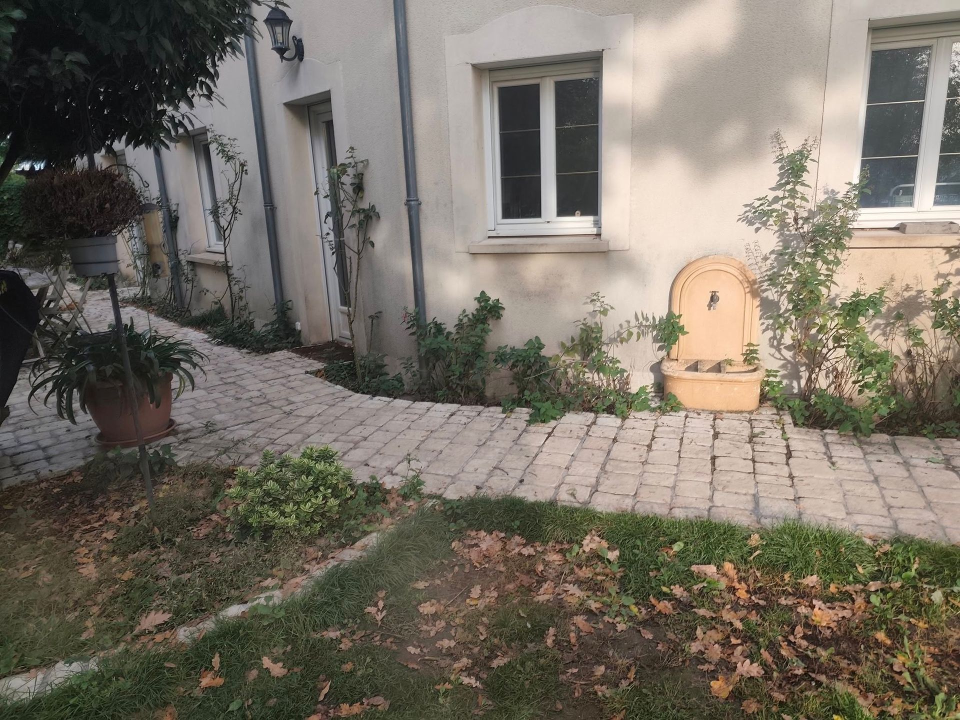 Un chemin pavé mène à un bâtiment doté de fenêtres et d'une fontaine murale décorative, le tout dans un cadre de jardin.