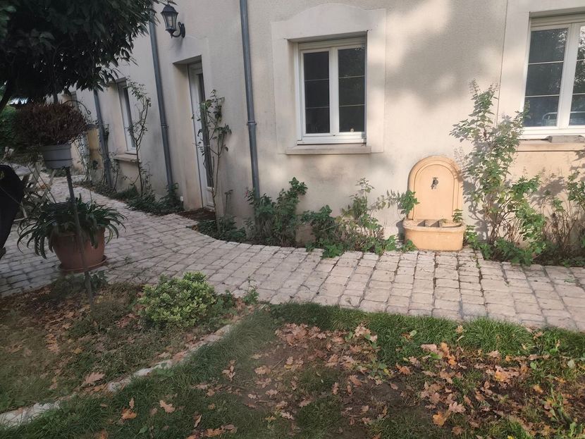 Un chemin pavé mène à un bâtiment de couleur claire avec une fontaine murale et des plantes en pot dans un jardin.