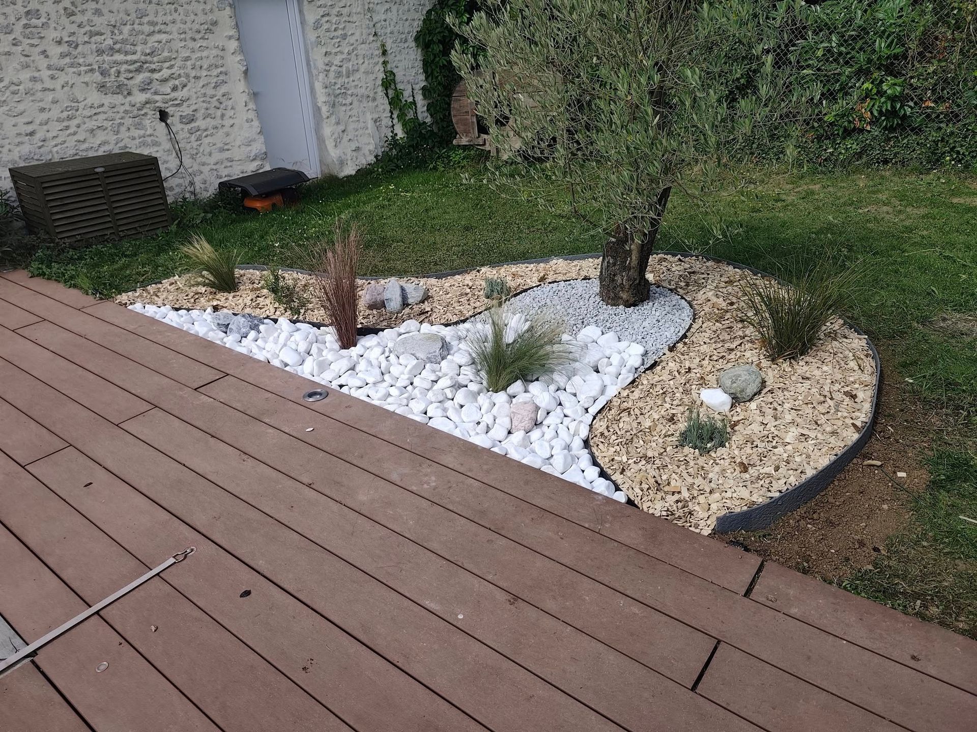Un parterre de jardin aménagé avec du gravier, des petites pierres et un olivier central, jouxtant une terrasse en bois.