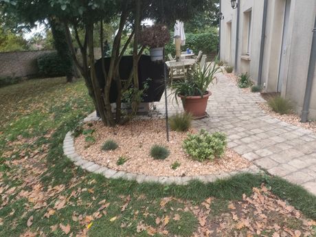 Une terrasse en pierre borde un parterre de jardin incurvé recouvert de gravier, agrémenté de petits arbustes et d'une plante en pot sous un arbre feuillu.