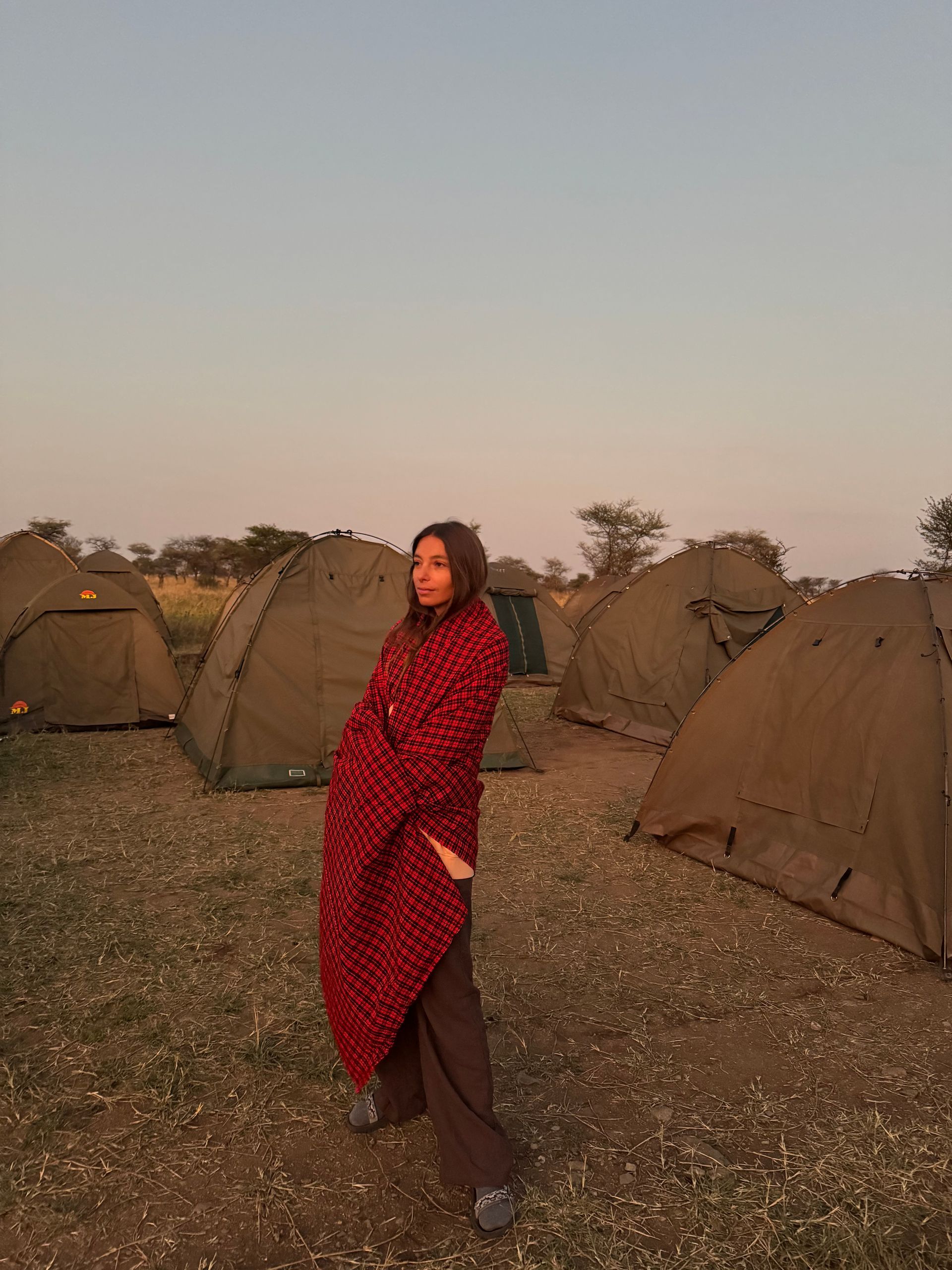 Una persona envuelta en una manta roja estampada está de pie cerca de tiendas de campaña en un campo al atardecer.
