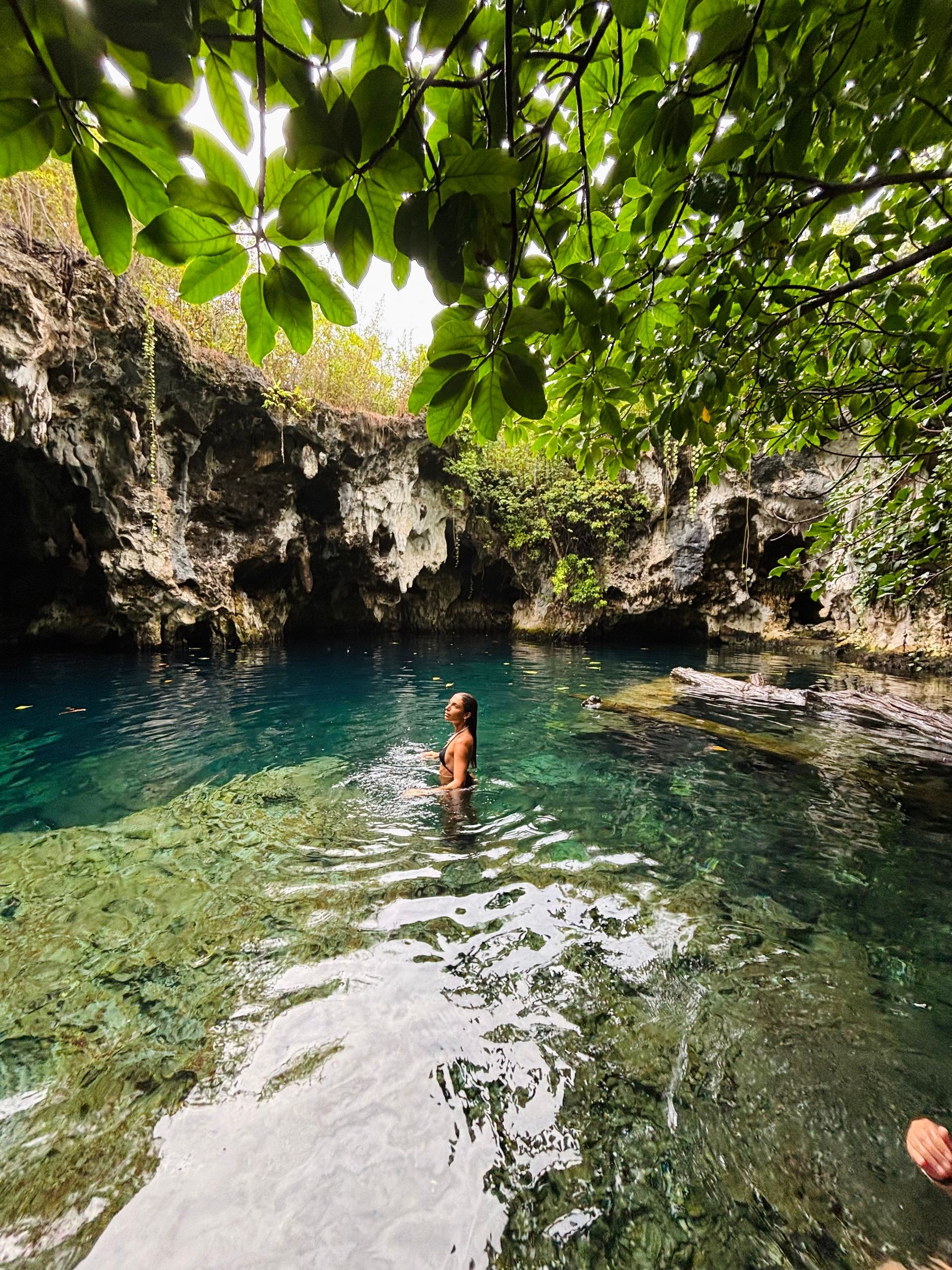 Persona de pie en agua cristalina de color turquesa, rodeada de acantilados rocosos y exuberante vegetación verde.