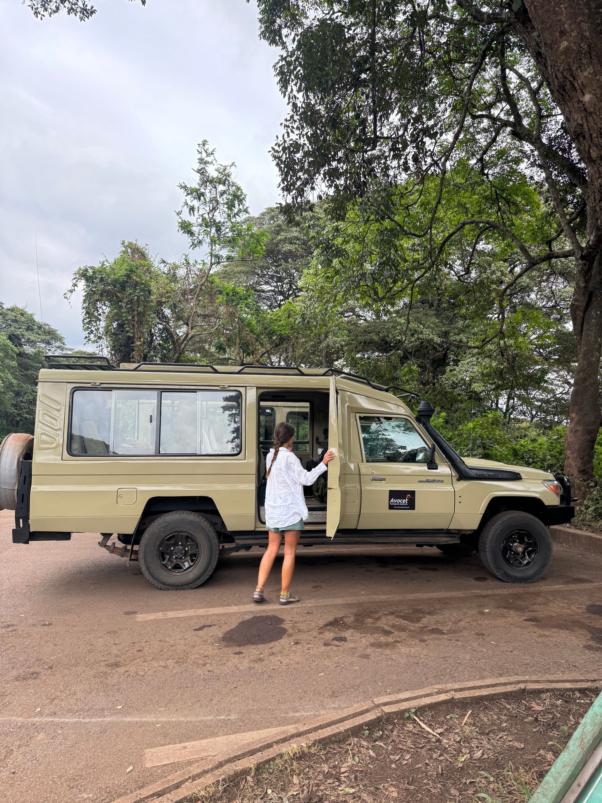 Una mujer abre la puerta de un vehículo de safari en una zona boscosa; vehículo color canela, neumáticos negros