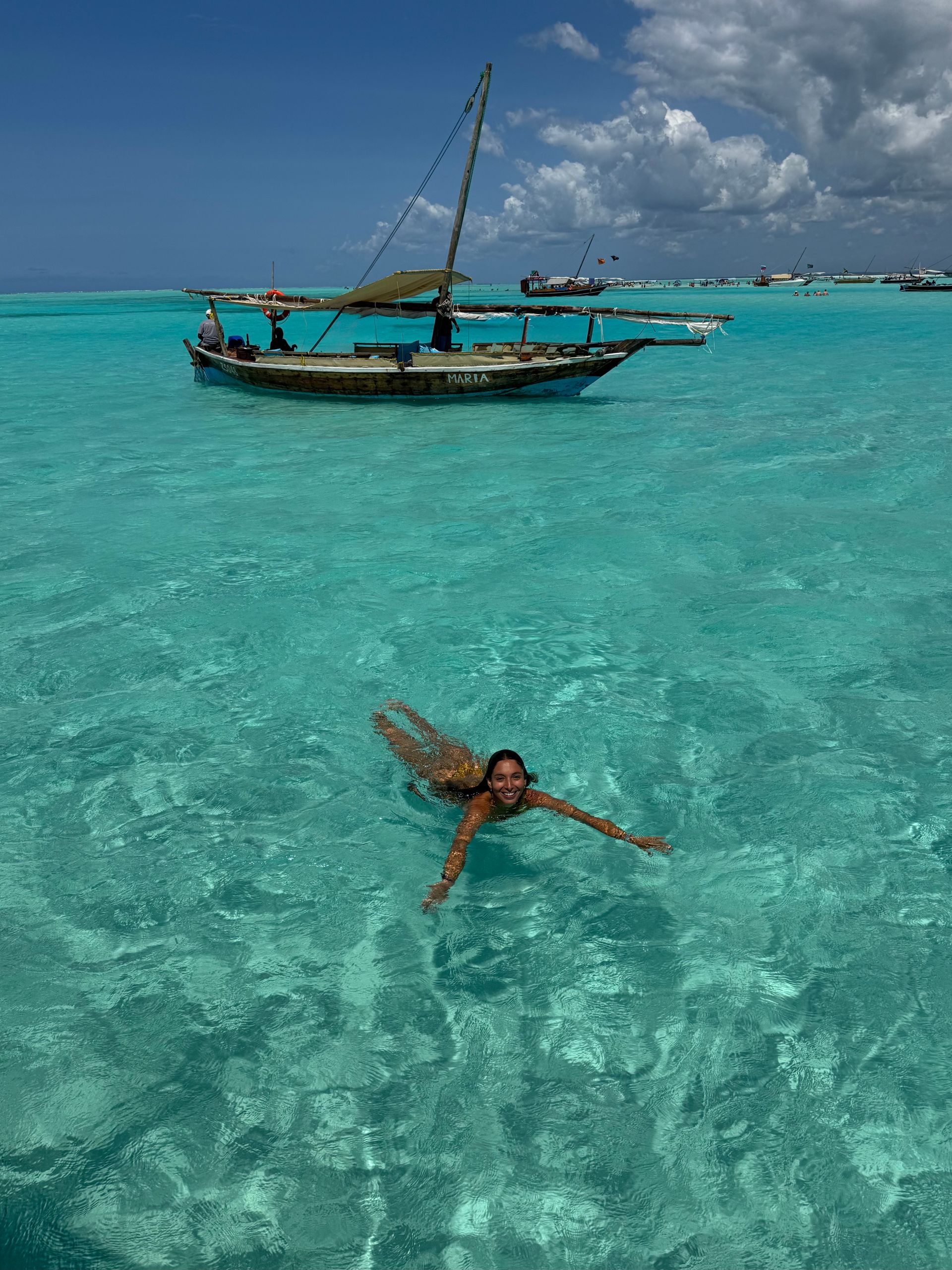 Una mujer nada en aguas turquesas con un barco al fondo en un día soleado.