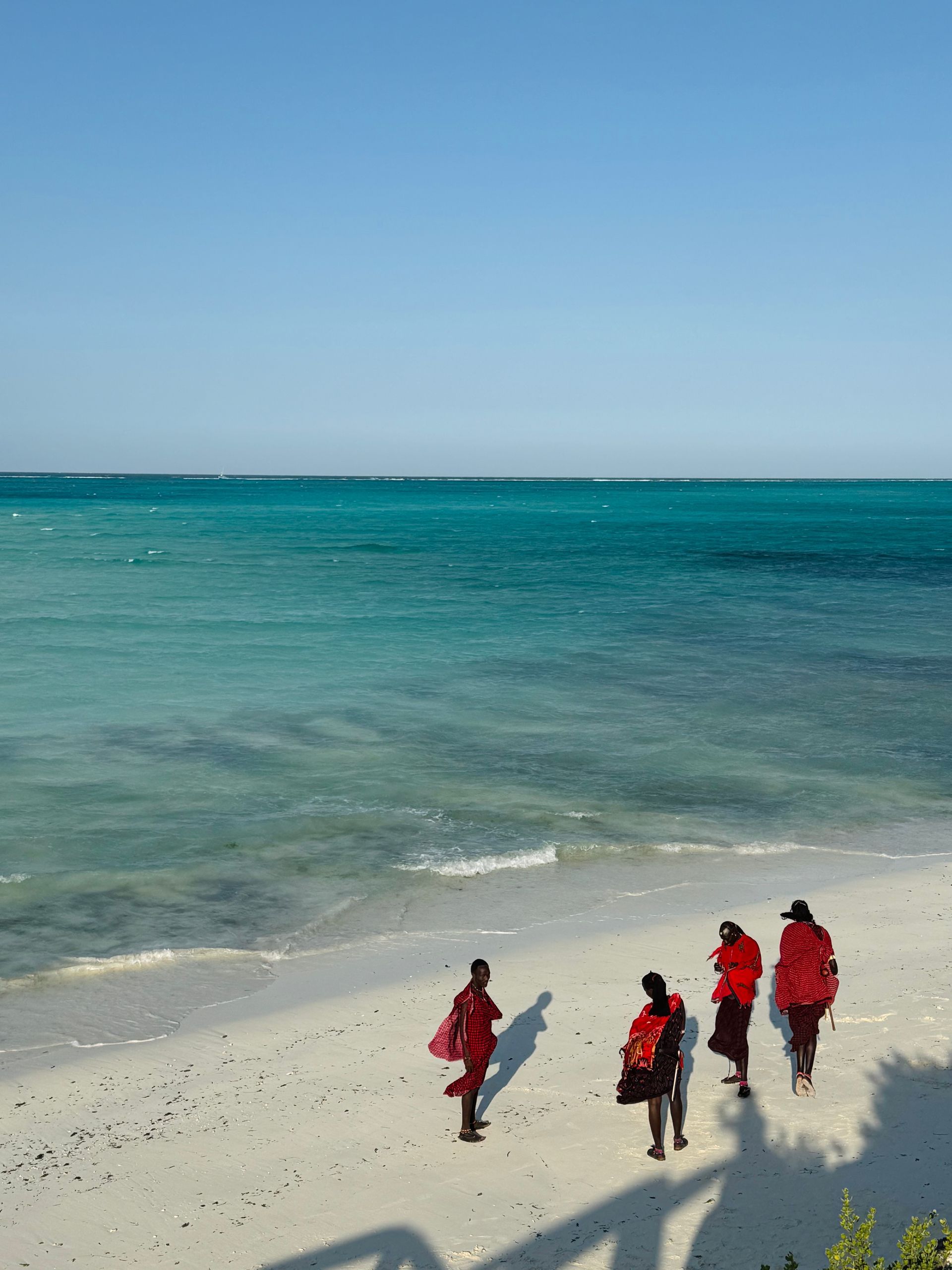 Personas vestidas de rojo caminan por una playa de arena blanca hacia aguas turquesas bajo un cielo azul.