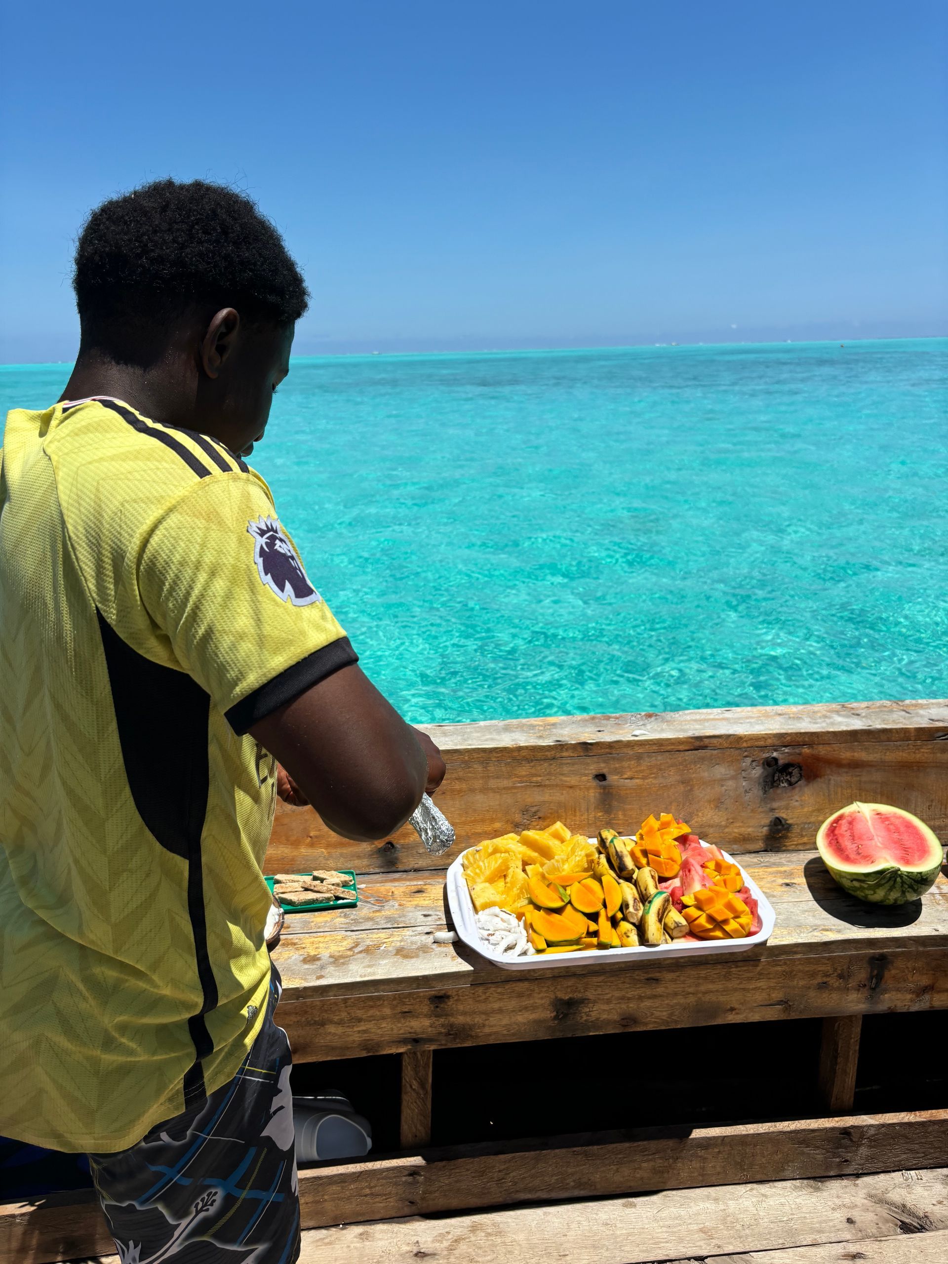 Hombre preparando fruta sobre una mesa de madera, con el océano turquesa de fondo.