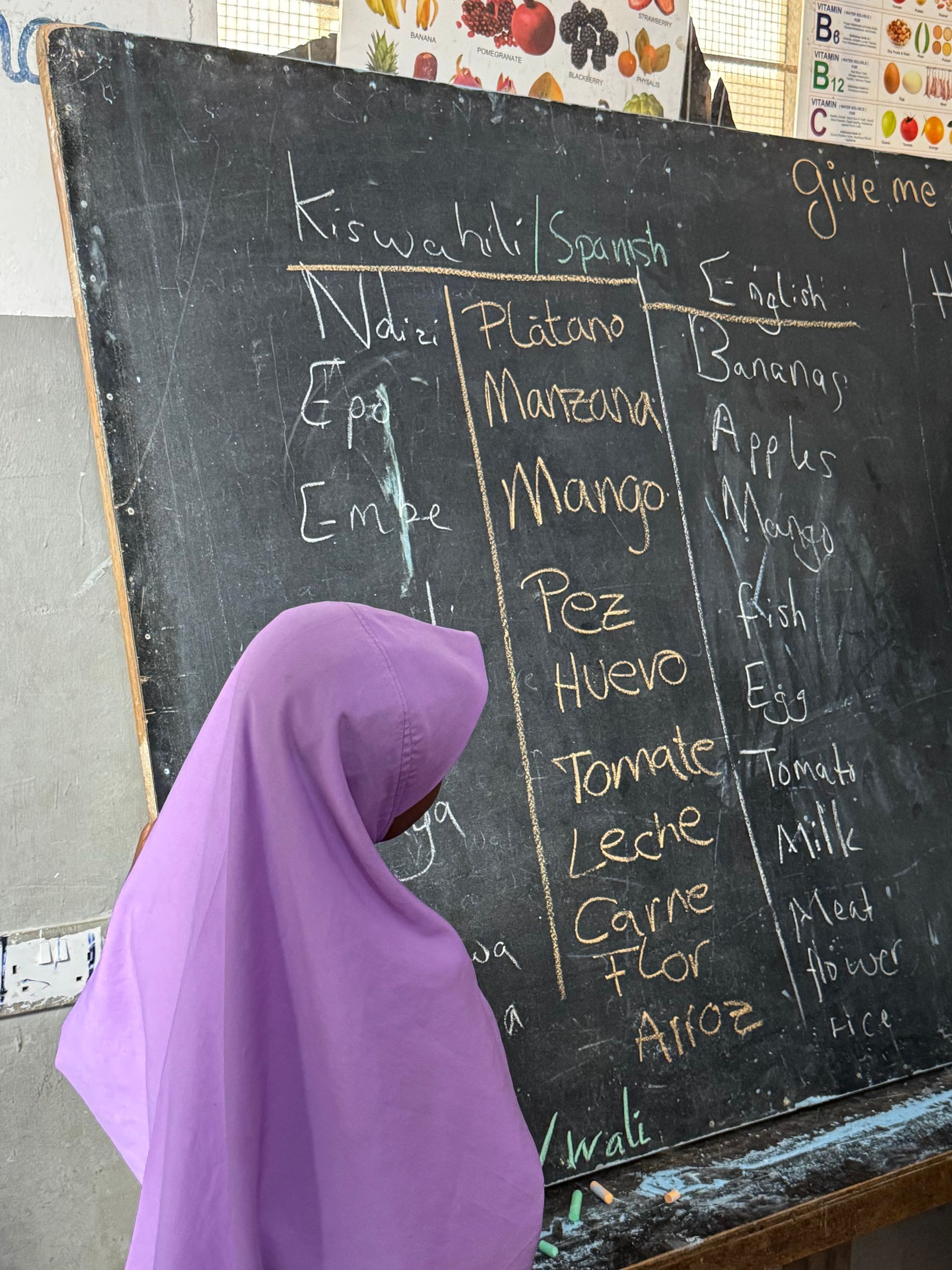 Una persona con hiyab morado mira una pizarra con nombres de comidas escritos en varios idiomas.