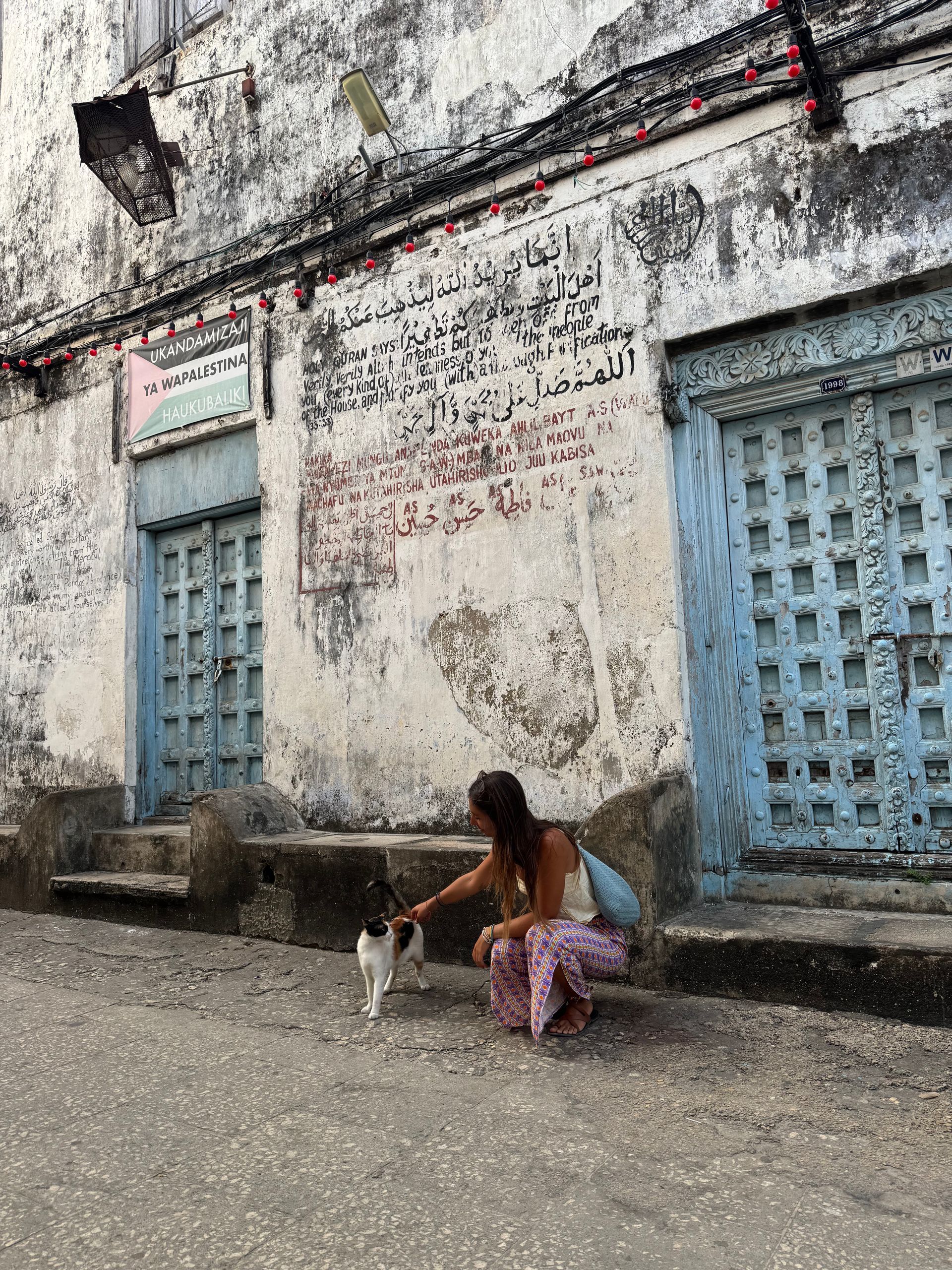 Una mujer acaricia a un pequeño gato en una calle con puertas antiguas de color azul claro y una pared descolorida