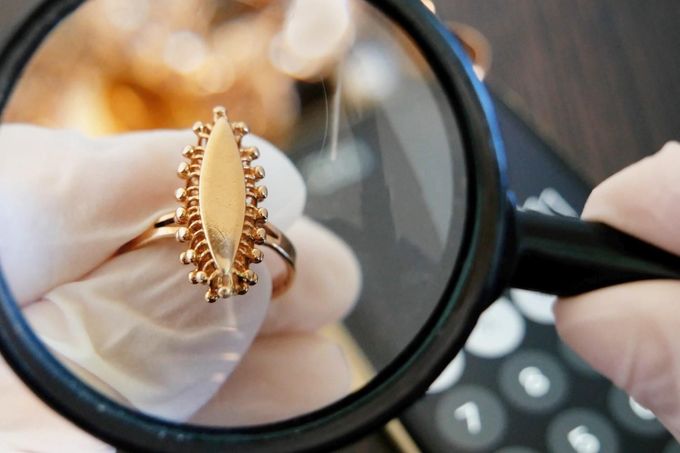 Unas manos enguantadas inspeccionan un anillo de oro con una lupa junto a una calculadora y documentos.