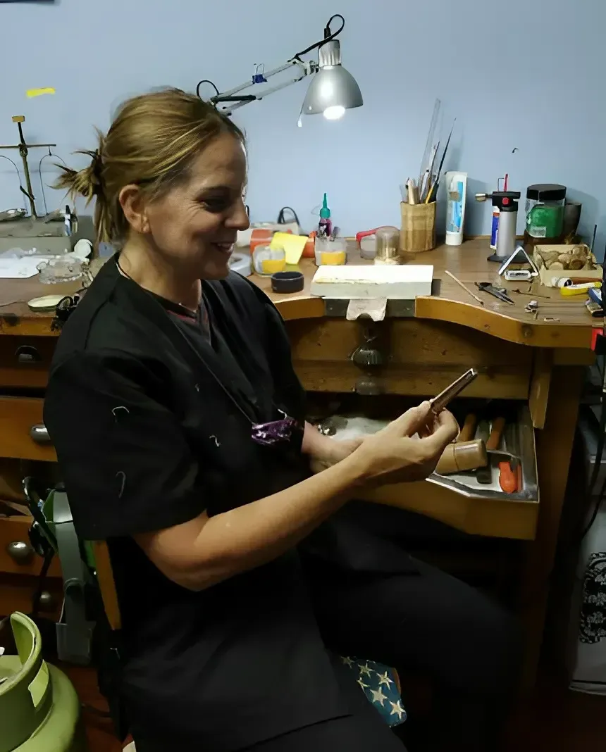 Persona sentada en un banco de trabajo desordenado, sosteniendo un teléfono bajo una lámpara de escritorio en un taller.