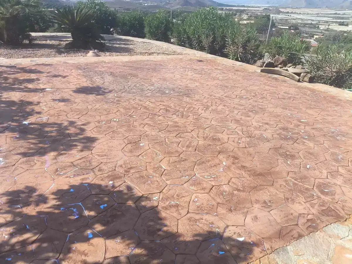 Patio de hormigón estampado de color marrón rojizo, con un patrón de formas irregulares; en un entorno ajardinado con plantas.