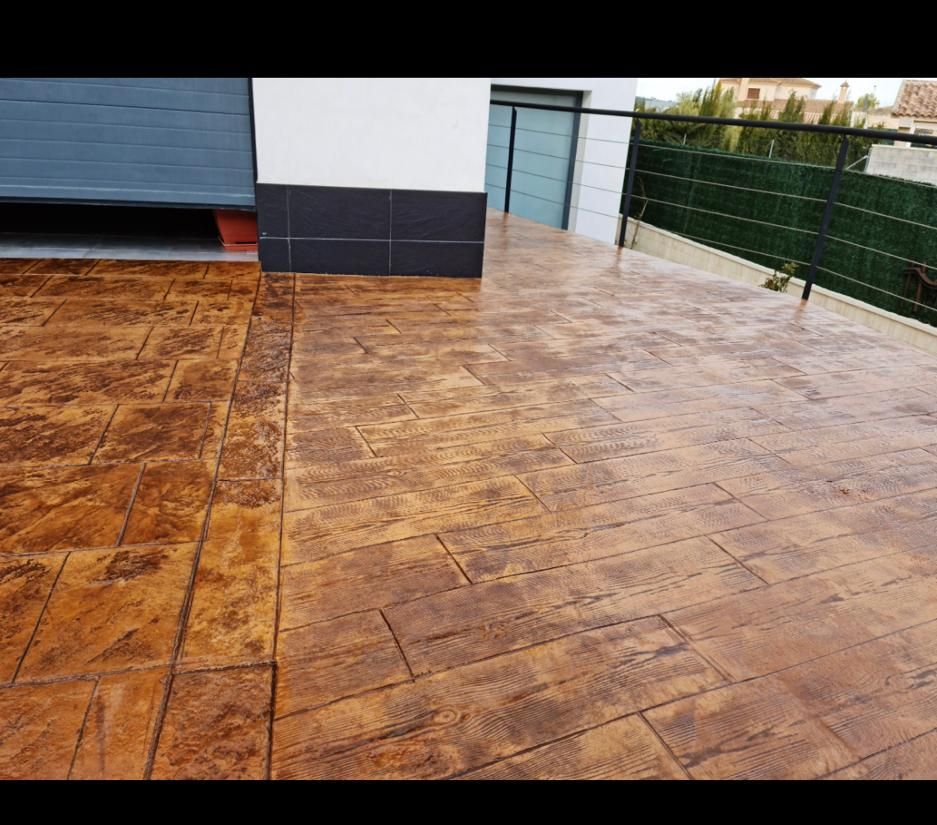Patio de hormigón estampado, marrón con patrón de ladrillo, junto a un edificio con puerta de garaje azul.