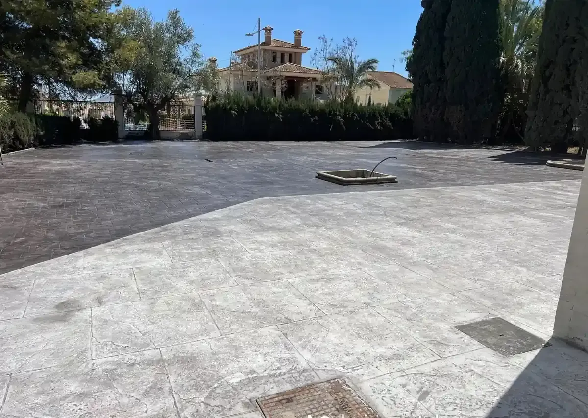 Patio de hormigón vacío con una zona oscura de grava y un edificio al fondo. Día soleado.