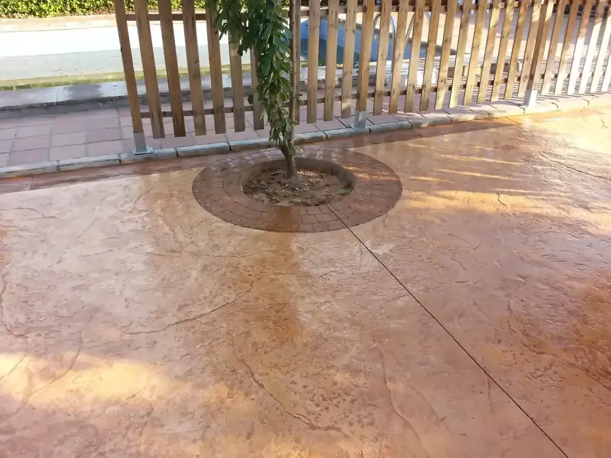 Patio de hormigón con un árbol, bordeado por una valla de madera, tonos marrones.