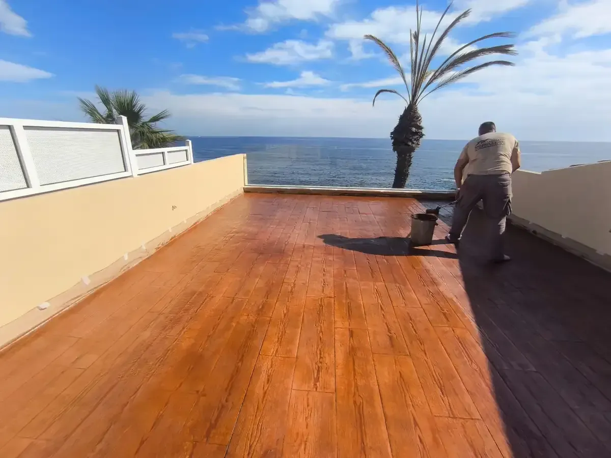 Hombre pintando una terraza de madera con vistas al océano con una palmera. Día soleado y radiante.