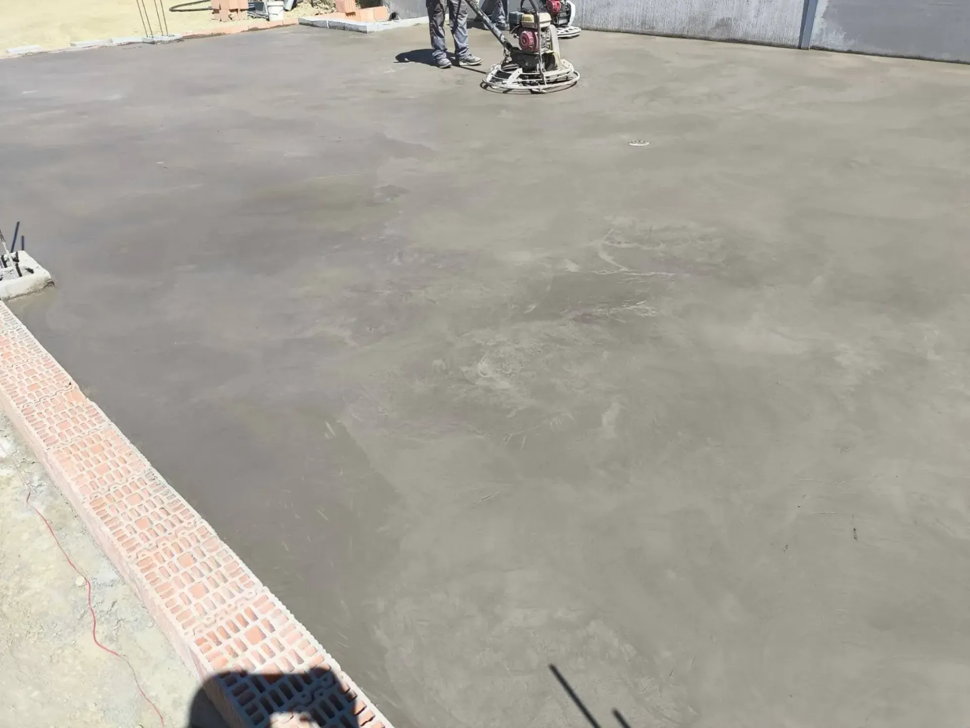 Obreros utilizan una allanadora mecánica para alisar hormigón húmedo sobre una gran superficie gris al aire libre.