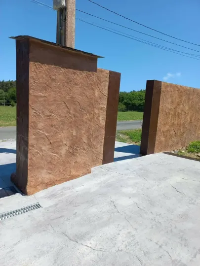 Dos pilares de hormigón marrón con superficie texturizada, situados en una zona de hormigón gris, bajo un cielo azul brillante.