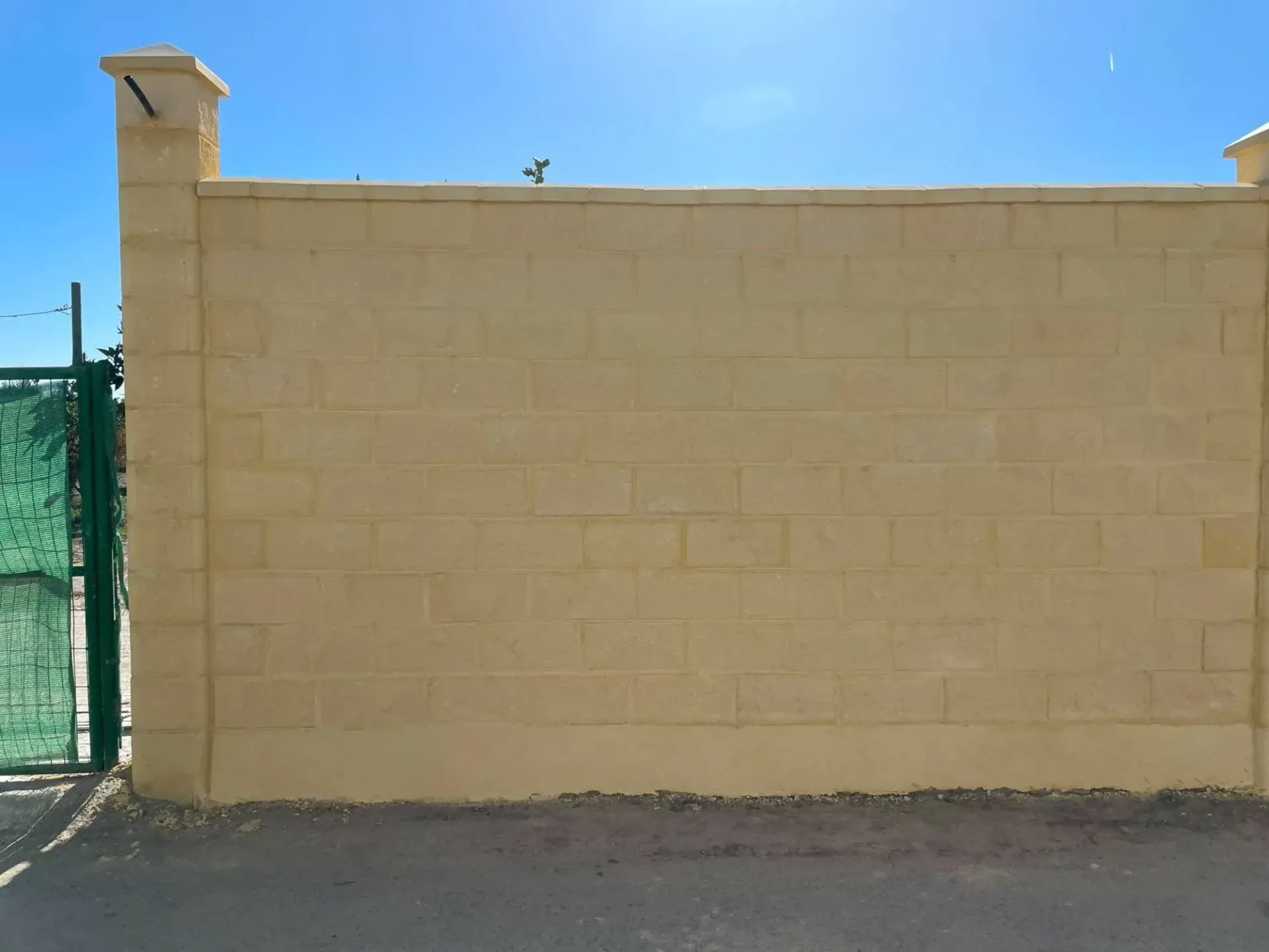 Muro de ladrillo amarillo con dos pilares decorativos, puerta verde a la izquierda, cielo azul arriba.